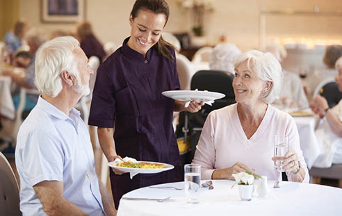 Senior Couple Being Served With Meal By Carer In Dining Room Of Retirement Home